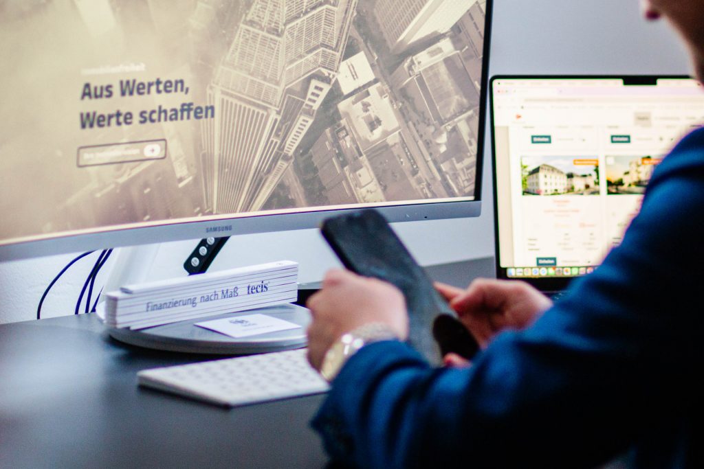 Mitarbeiter am Schreibtisch bei geöffneter Webseite im Hintergrund und einem Handy in der Hand